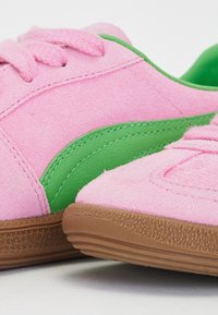 Pink suede sneakers with green side stripes and brown textured rubber soles, featuring thick pink laces, resting on a white surface.