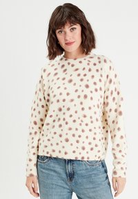Pull crème à pois marron, col rond, avec des poignets et un ourlet côtelés ; porté avec un jean en denim bleu clair. Texture douce, coupe décontractée.