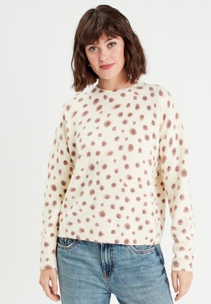 Crème trui met bruine stippen, ronde hals, geribde manchetten en zoom; gedragen met lichtblauwe denim jeans. Zachte textuur, ontspannen pasvorm.