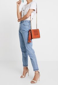 Pomarańczowa skórzana torebka o plecionej fakturze z ozdobnym chwostem. Noszona z koszulką z nadrukiem i niebieskimi dżinsami z wysokim stanem, uzupełniona beżowymi szpilkami.