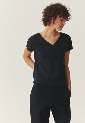 Mujer con cabello corto y rizado, vestida con una camiseta negra de mangas cortas y escote en V, y pantalones negros, de pie con una mano detrás de la espalda.