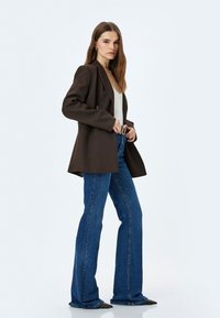 Blazer marrone su una maglia chiara, abbinato a jeans blu a zampa. L'outfit presenta una vita stretta e scarpe nere a punta.