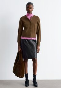 Braune Strickjacke, die vorne zugeknöpft ist, über einem pinken Rollkragenpullover, kombiniert mit einem schwarzen Rock im Krokodilmuster und schwarzen Stiefeletten mit Absatz. Braune Fellhandtasche.