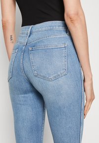 Jeans de denim azul claro con un diseño ajustado, que cuentan con bolsillos traseros y costuras en contraste a lo largo de las costuras. Textura suave.