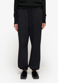 Czarne dresy z elastycznym paskiem w talii, sznurkiem i ściągaczami u dołu nogawek. Zawierają małe wyszywane logo na lewym biodrze.