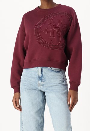 Frau trägt einen burgunderfarbenen Sweatshirt mit Rundhalsausschnitt und geprägt gelagertem Logo-Design kombiniert mit hellblauen Jeans mit hoher Taille, steht vor weißem Hintergrund.