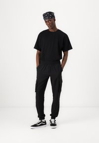 Svart T-skjorte med korte ermer, svarte cargo-bukser med sidelommer og svarte sneakers med hvite detaljer. Bandana med hvitt mønster.