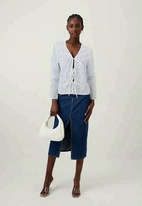 Λευκό πουκάμισο με βολάν και μακριά μανίκια, με κορδόνι στο μπροστινό μέρος, συνδυασμένο με τζιν φούστα σε μπλε απόχρωση που διαθέτει σκίσιμο μπροστά. Το μοντέλο κρατά μια λευκή τσάντα.