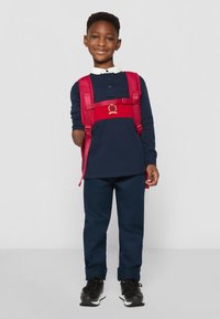 Granatowa koszula polo z długim rękawem i białym kołnierzykiem, granatowe spodnie, czerwony plecak z złotym emblematem oraz czarne sneakersy.