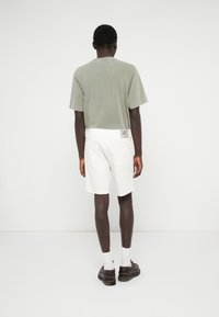 Világos zöld pamut póló fehér rövidnadrággal; a rövidnadrág hátsó zsebében címke található. Barna cipő és fehér zokni teszi teljessé a megjelenést.