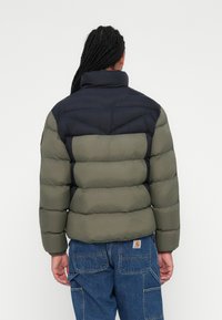 Puffjakke i olivengrøn og navy. Har en høj krave, quiltet design og elastiske manchetter. Båret med blå jeans, der viser en afslappet pasform.