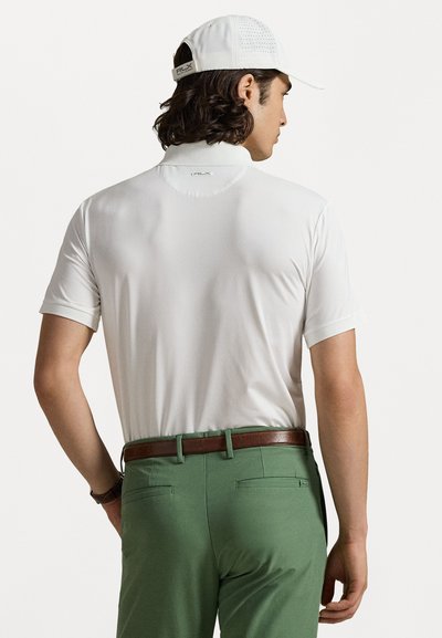 Vit kortärmad polo med en ståkrage, i kombination med gröna byxor och ett brunt bälte, visad bakifrån.