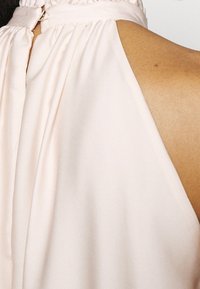 Primer plano del hombro y la parte superior de la espalda de una mujer que lleva una blusa sin mangas de color rosa pálido con un escote fruncido y cierre de botón dorado.