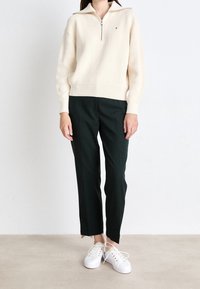 Kobieta ubrana w kremowy sweter zapinany na zamek, czarne spodnie o prostym kroju i białe trampki, stojąca na gładkiej podłodze na białym tle.