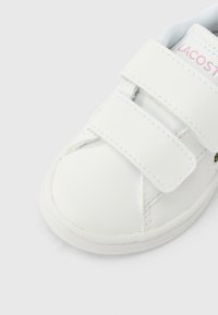 Baskets blanches avec bandes Velcro, dessus synthétique lisse, bout arrondi et détail discret du logo en rose. Semelle texturée pour une bonne adhérence.