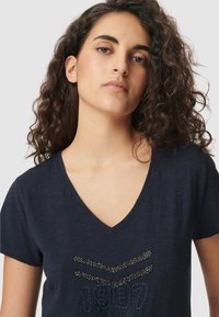 Joven mujer con cabello rizado oscuro que lleva una camiseta de cuello en V color marino con chevrones de cuentas y "1987" bordado en el frontal.