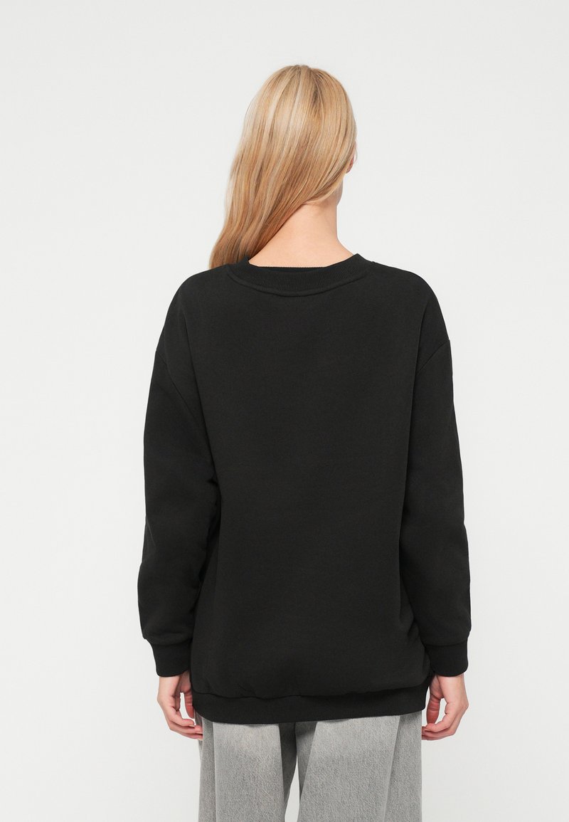 Blond persoon in een losse zwarte sweatshirt en lichtgrijze broek, van achteren gezien tegen een effen witte achtergrond.