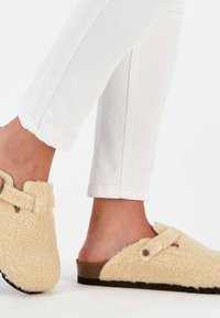 Fuzzy beige clogs with a textured finish, featuring a brown accent strap, a closed toe, and a low cork-like sole, worn with white jeans.