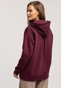 Bordowy hoodie o miękkiej fakturze, z szerokim dekoltem i dużym kapturem. Projekt ma luźny krój, zaprezentowany z beżowymi spodniami z korduroju.