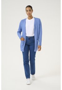 Cardigan bleu clair avec manches longues et poches frontales, superposé sur un t-shirt blanc, associé à un jean bleu et des baskets blanches.