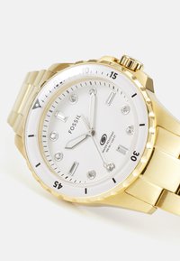 Montre Fossil en ton or avec un cadran blanc, des accents en cristal, un bracelet en acier inoxydable et une lunette rotative. Résistante à l'eau jusqu'à 100 mètres.