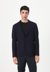 Mornarški blazer z oblikovano zasnovo, dvema sprednjima žepoma, reberastimi reverji in dvema črnima gumboma. Nosi se čez črno srajco s gladko teksturo.