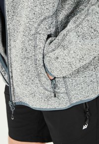 Grå mönstrad zip-up jacka med fleece-textur, med mörkblå detaljer, handfickor och justerbara dragsnören i kant.