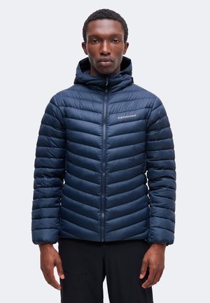 Homme portant une veste matelassée à capuche bleu marine, zippée, debout devant un fond clair uni.