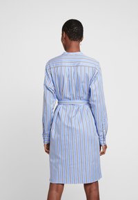 Personne avec les cheveux courts portant une robe chemise ceinturée à rayures bleues et blanches, se tenant de dos contre un fond uni.