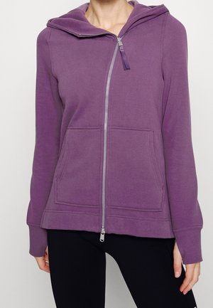 Femme portant un sweat à capuche violet zippé avec poches avant et manches longues, associé à un pantalon noir sur fond blanc.