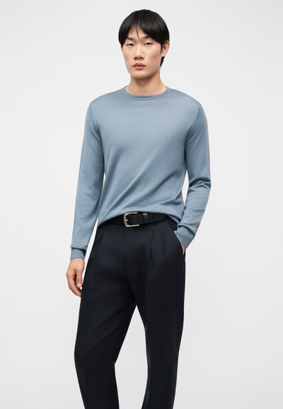 Νεαρός άνδρας στέκεται όρθιος, φορώντας ένα ανοιχτό γαλάζιο πουκάμισο με μακριά μανίκια, που έχει μπει μέσα σε μαύρο παντελόνι με μαύρη ζώνη, με το ένα χέρι στην τσέπη.