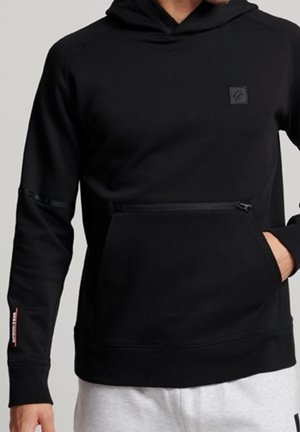 Homme portant un sweat à capuche noir avec des poches zippées à l'avant et aux bras, debout devant un fond gris uni.