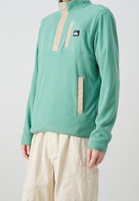 Pullover in pile verde chiaro con collo a mezza zip, patta con bottoni beige e tasca laterale. Presenta un piccolo logo sul petto.
