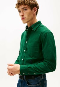 Groene overhemd gemaakt van katoen, met een borstvak en geborduurd logo aan de onderzijde. Draagt bij blauwe spijkerbroeken en een zwarte riem.