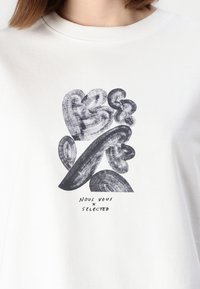Hvit bomull T-skjorte med et svart abstrakt grafisk design med svingende former og tekst som sier "NOUS VOUS X SELECTED" under.
