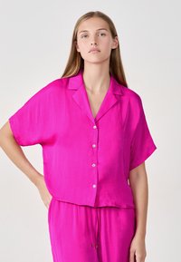 Camisa fucsia de manga corta con botones, hecha de una tela ligera, que presenta un cuello clásico y un corte holgado. Textura suave con un ligero brillo.