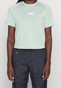 Camiseta corta de color verde claro con mangas cortas, con un pequeño logo blanco en el pecho. Combinada con pantalones oscuros y ligeros con cintura elástica.