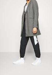 Grijze lange blazer over een wit T-shirt, gecombineerd met zwarte broek met een wit accent en aangetrokken enkels, en witte hoge sneakers.