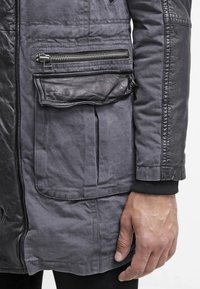 Grå jacka med en blandning av strukturerade material. Inkluderar en ficka med dragkedja, en stor framficka och ribbade ärmar.