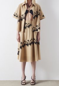 Beige shirt en rok set met zwarte abstracte patronen, met eronder een cropped top. Stof lijkt glad; stijl is relaxed. Zwarte sandalen met hakken.