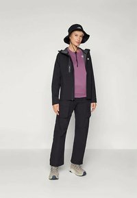 Ženska nosi črno vedro, črno jakno, vijolično majico, črne cargo hlače in bež pohodniške čevlje, stoji pred enobarvno ozadjem.