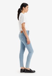 Mujer con coleta oscura, vistiendo una camiseta blanca, jeans ajustados de mezclilla azul claros con dobladillo sin terminar y mocasines negros, de pie de perfil sobre un fondo blanco.