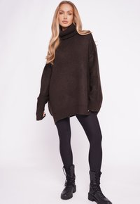 Übergroßer brauner Strickrollkragenpullover mit einer strukturierten Oberfläche, kombiniert mit schwarzen Leggings und knöchelhohen schwarzen Schnürstiefeln.