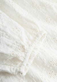 Witte stof met geborduurd bloemrijk oogjespatroon en elastische geplooide stiknaden op een naad of manchet.