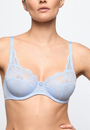 Frau trägt einen hellblauen Spitzen-Bügel-BH mit geometrischen Mustern und verstellbaren Trägern, gezeigt von den Schultern bis zur Mitte des Bauches.