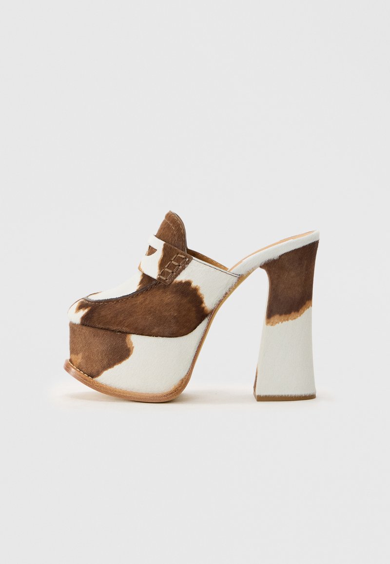 Brūnas un baltas govs ādas platformē iešļūcenes ar masīvu augstu papēdi un loafers stila augšdaļu uz vienkārša balta fona.