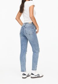 Jean en denim bleu taille haute avec une finition délavée, légère usure aux genoux, cinq poches et coupe slim. Associé à des baskets blanches.