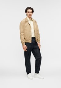 Καφέ μπουφάν bomber πάνω από ένα ανοιχτό πουκάμισο με γιακά, σε συνδυασμό με μαύρο ελαστικό παντελόνι και λευκά αθλητικά παπούτσια. Απλός σχεδιασμός με ομαλές υφές.