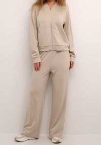 Ensemble jogger beige en tissu doux et extensible. Comprend une veste zippée, une coupe décontractée et une taille élastique sur le pantalon. Agrémenté de poches.