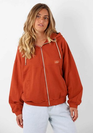 Haut à zip couleur rouge brique avec une poche avant, cordons de serrage et patch logo. Tissu doux, coupe ample et poignets ribbées avec ceinture.